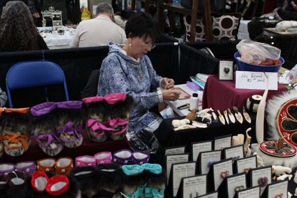 An Alaska Native artist sits behind a table covered in their work at an art fair. 