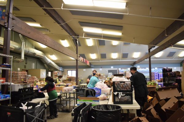 Workers sort crates of food at large tables inside a large room with skylights.