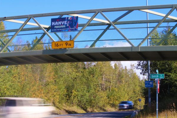 A blue Kanye sign hangs on an elevated footbridge as two cars pass