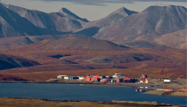 A few small buildings and gravel pits are seen from across blue water below mountains