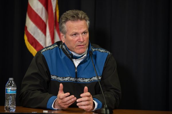 A white man speaks as he sits at a table with an american flag in the background