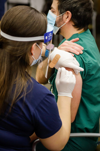 a person wearing a face shield and mask administers a shot to someone wearing scrubs and a mask