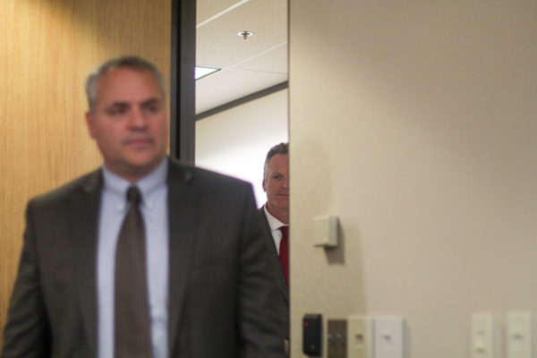 Governor Dunleavy peeks through a door with a man in a suit in the foreground