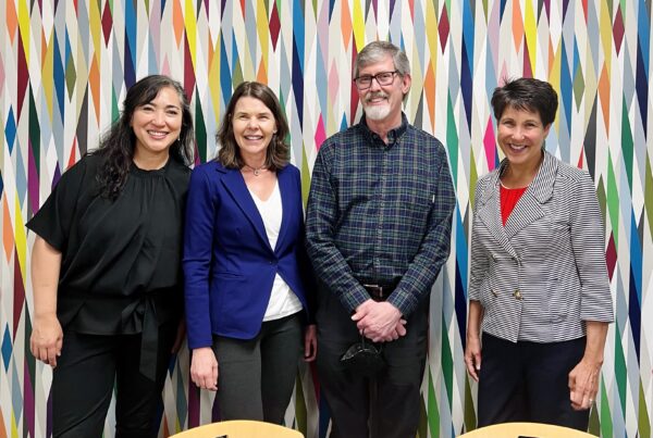 Four people stand together smiling and posing for the camera in front of a multicolored background.