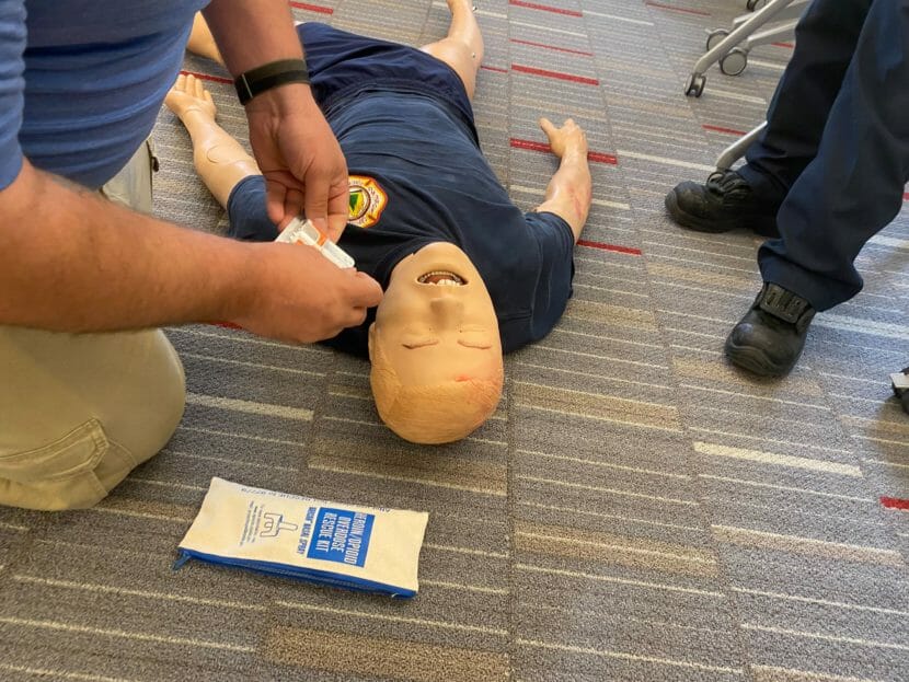 Interior: a CPR dummy rests on the ground as volunteers prepare to practice administering Narcan.